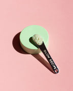 Glow Recipe silver skin care spatula with cream in bowl n top of green stand with pink background.