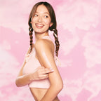 Woman wiping cream Watermelon Glow pink product on arm. She is standing against a pink background.