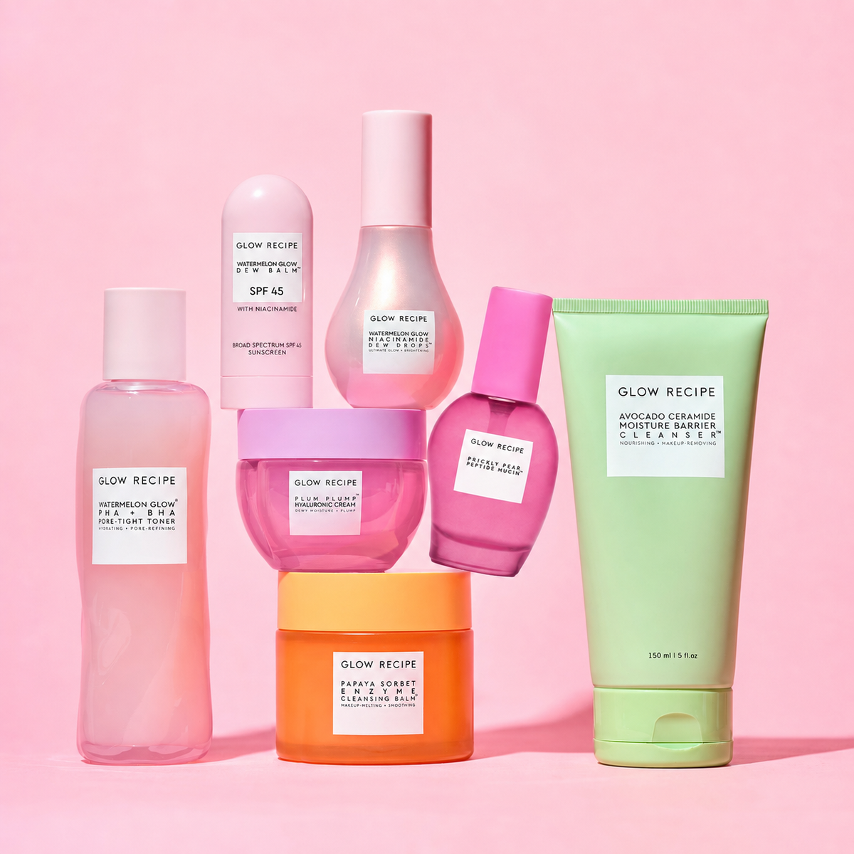 Six Glow Recipe skincare products in various shapes and colors (pink, purple, orange, and light green) arranged together on a solid pink background.